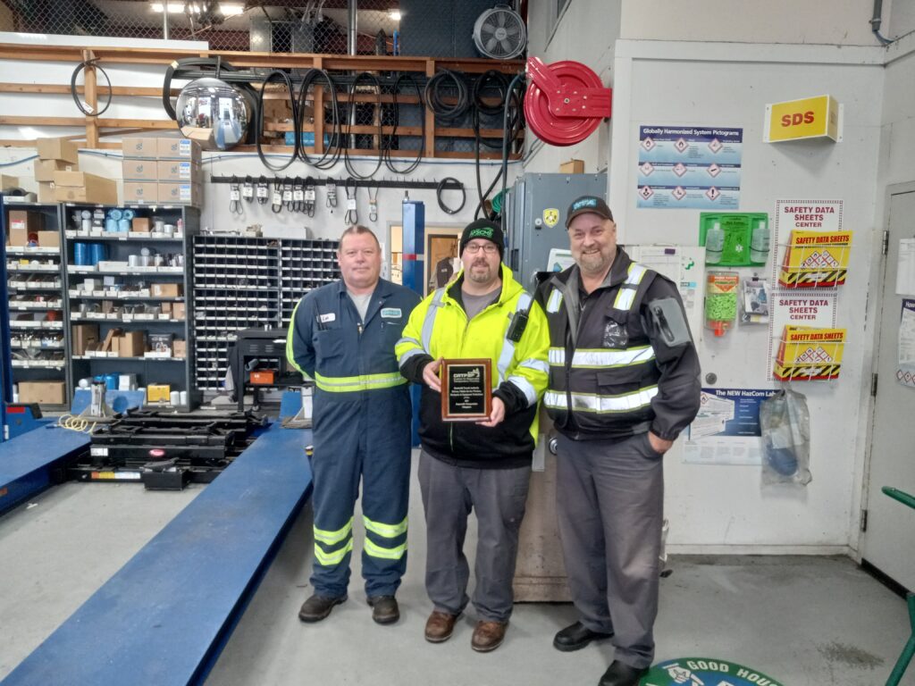 Three Humboldt Transit Authority employees in the shop, the person in the center holding a plaque