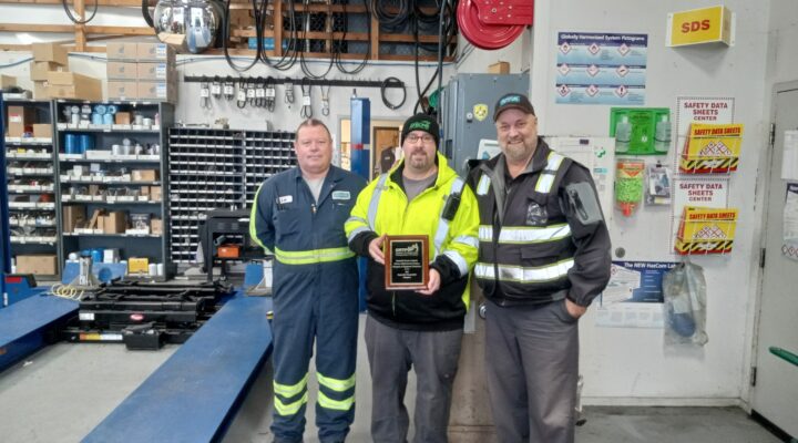 Three Humboldt Transit Authority employees in the shop, the person in the center holding a plaque