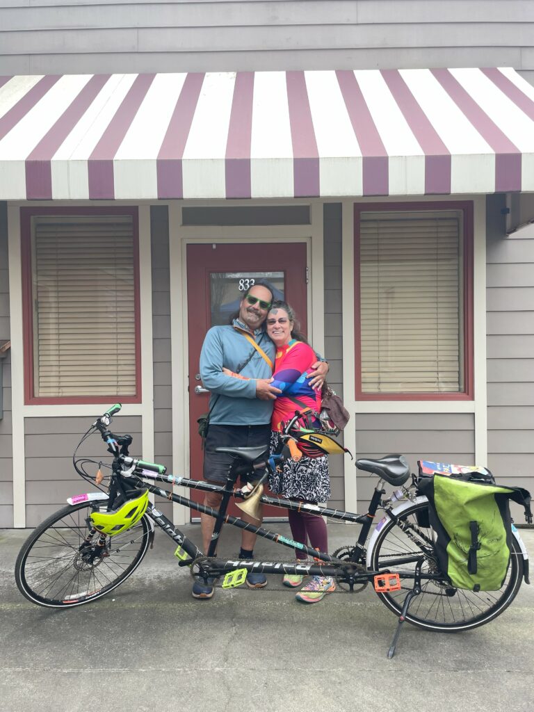 Two people hug while standing behind a tandem bicycle and in front of a grey and red building with a striped awning