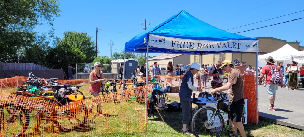 A grassy area with bikes is enclosed by orange temporary fencing and a canopy that reads "Free Bike Valet," while a volunteer checks in a bike