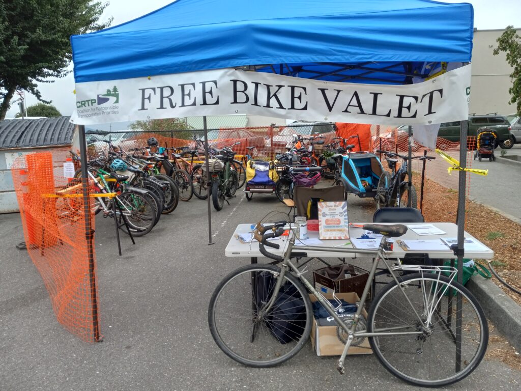 A bicycle leans against a table under an awning with a sign that reads "Free Bike Valet." Behind the table many bikes are parked and enclosed in orange temporary fencing.