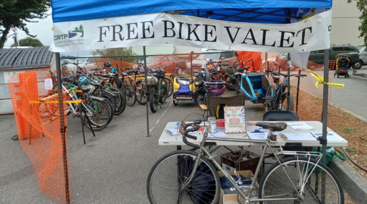 A bicycle leans against a table under an awning with a sign that reads "Free Bike Valet." Behind the table many bikes are parked and enclosed in orange temporary fencing.