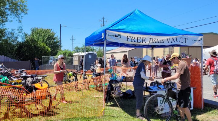 A grassy area with bikes is enclosed by orange temporary fencing and a canopy that reads "Free Bike Valet," while a volunteer checks in a bike