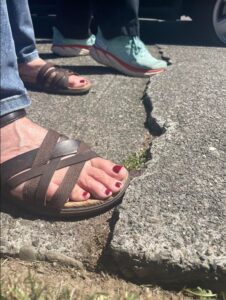 Two people's feet are shown, one in sandals with red-painted toenails, the other in athletic shoes. The toes of one foot of each person are catching on a crack in the sidewalk.