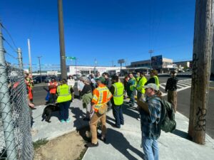 About twenty people, many wearing orange and yellow safety vests, stand on the sidewalk on a corner on 5th Street in Eureka