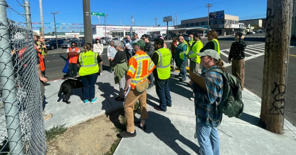 About twenty people, many wearing orange and yellow safety vests, stand on the sidewalk on a corner on 5th Street in Eureka