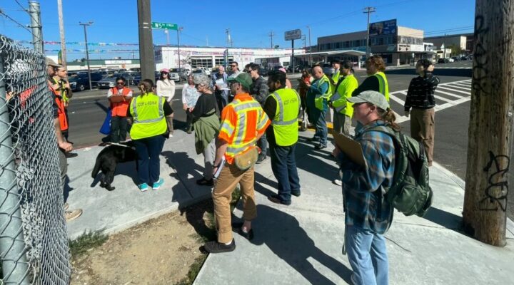 About twenty people, many wearing orange and yellow safety vests, stand on the sidewalk on a corner on 5th Street in Eureka