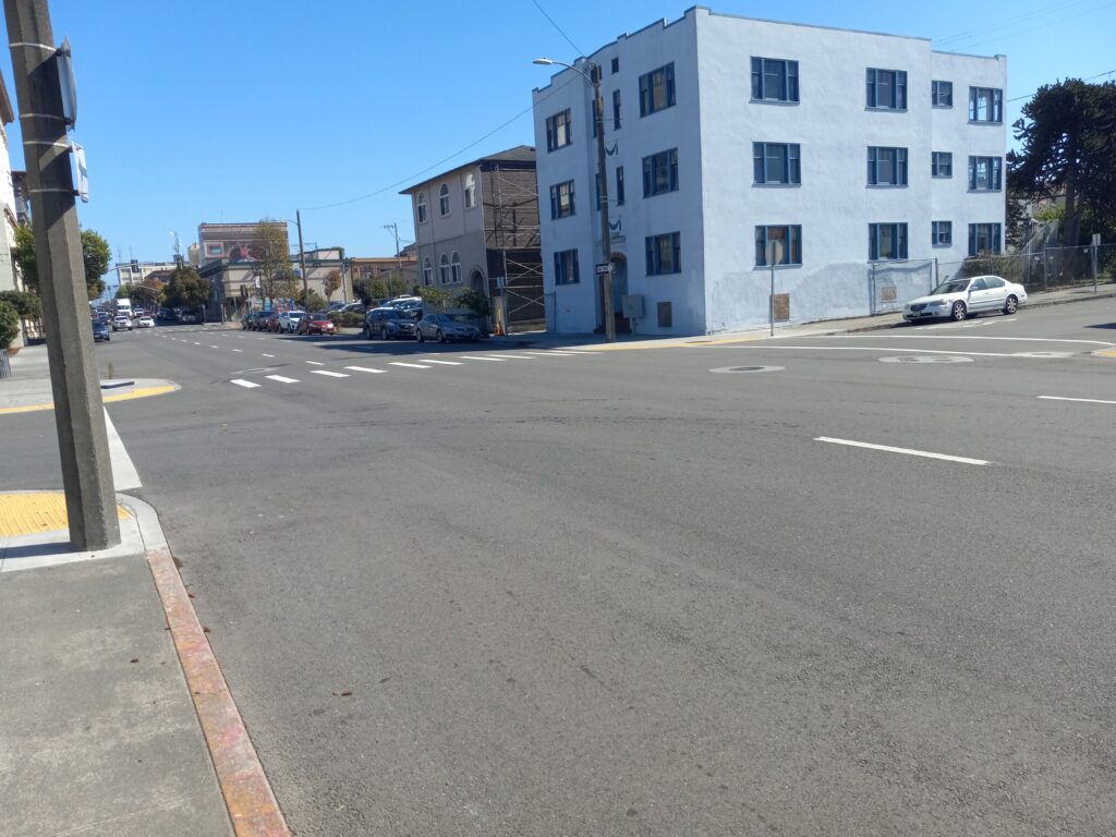 Three lanes of one-way traffic at an intersection. Crosswalk markings are visible at the far side of the intersection, but not the near side. A 3-story building is visible across the street.