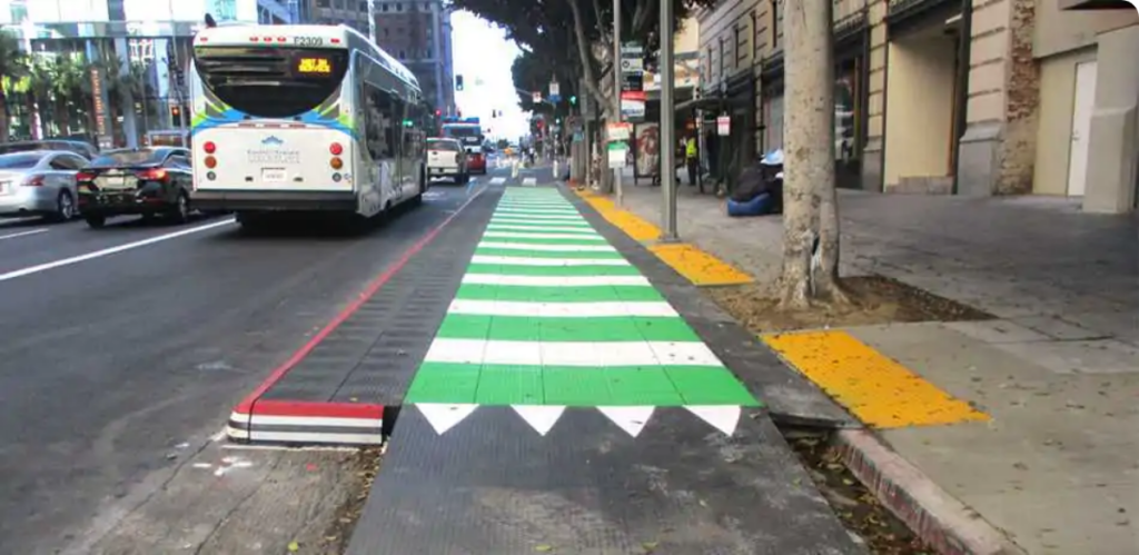 A raised platform extends a sidewalk into the street next to a bus. A ramp brings a green-painted bike lane up the level of the platform.
