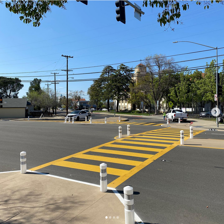 A yellow ladder-style crosswalks crosses a wide intersection. Plastic posts surround the ends of the crosswalk and refuge islands in the middle