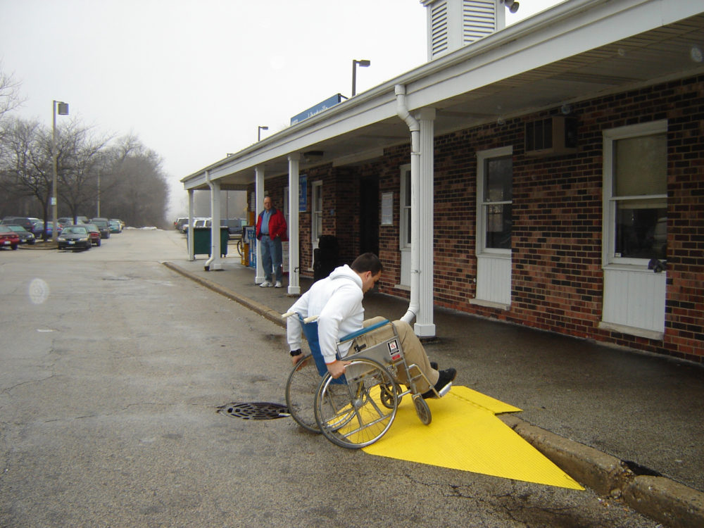 A man in a wheelchair wheels up a yellow curb ramp toward a row of apartments