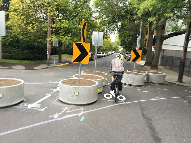 A person bikes between concrete planters with traffic signs in them on a tree-lined street