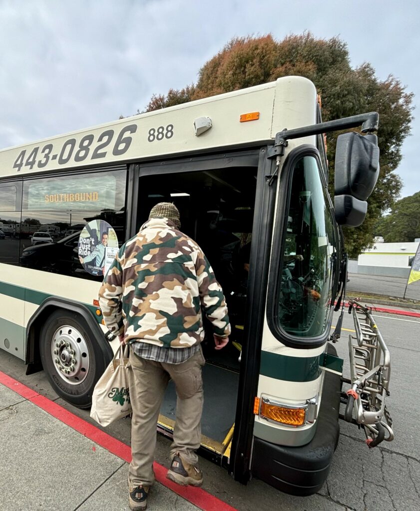 A person in tan pants and a camouflage patterned sweater steps onto a bus from the sidewalk.