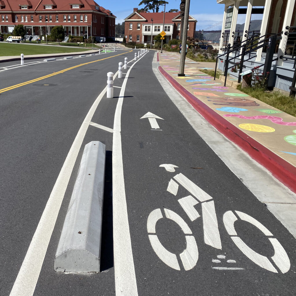 A bike lane with a bicycle stenciled in white paint is bordered by white-painted concrete curbs, plastic posts, and a brightly painted sidewalk