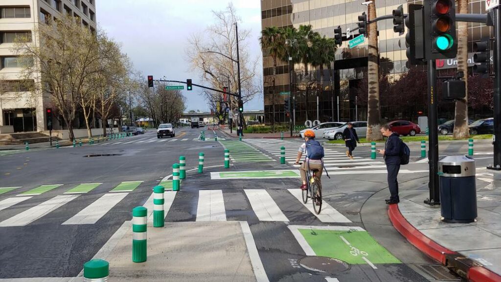 A large 4-way intersection with green-painted bike lanes along all streets, protected at the corners by green plastic posts