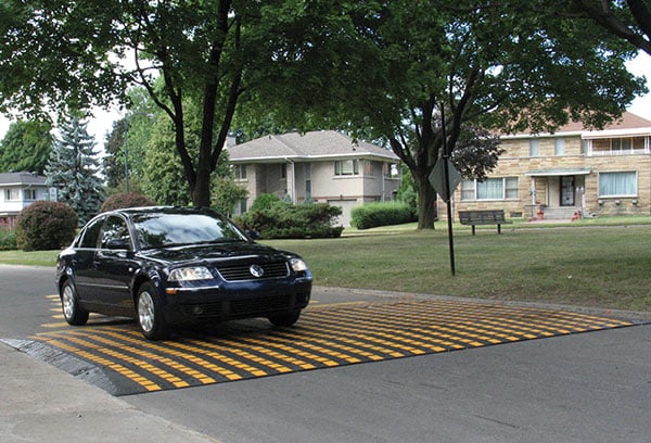 A black car on a residential tree-lined street drives over a plastic black-and-yellow speed table