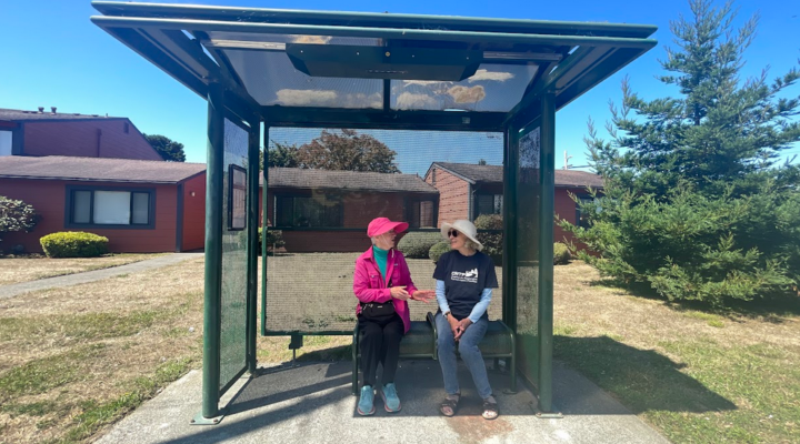 Two people sit and talk to each other on a bench in a bus shelter on a sunny day, one wearing a pink jacket and hat, the other a sun hat.