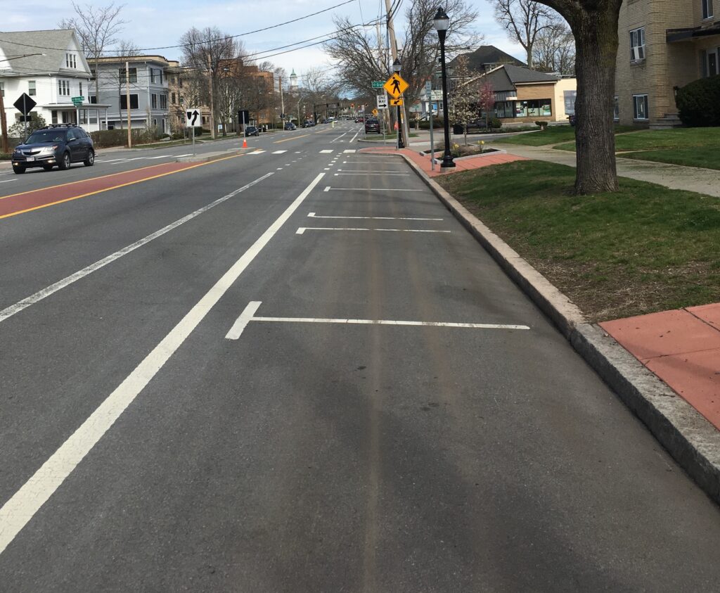 A street with a bike lane, a parking lane, a landscaped buffer and a sidewalk. The parking lane includes white paint marks to delineate individual spaces.