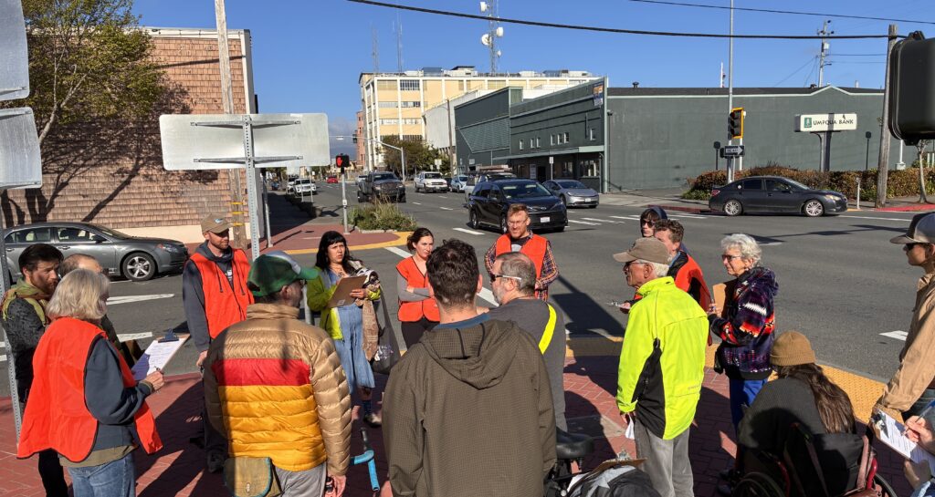 A group of people of varying ages, many wearing reflective orange safety vests, are gathered talking on a street corner. In the background, cars pass on a busy three-lane one-way street.