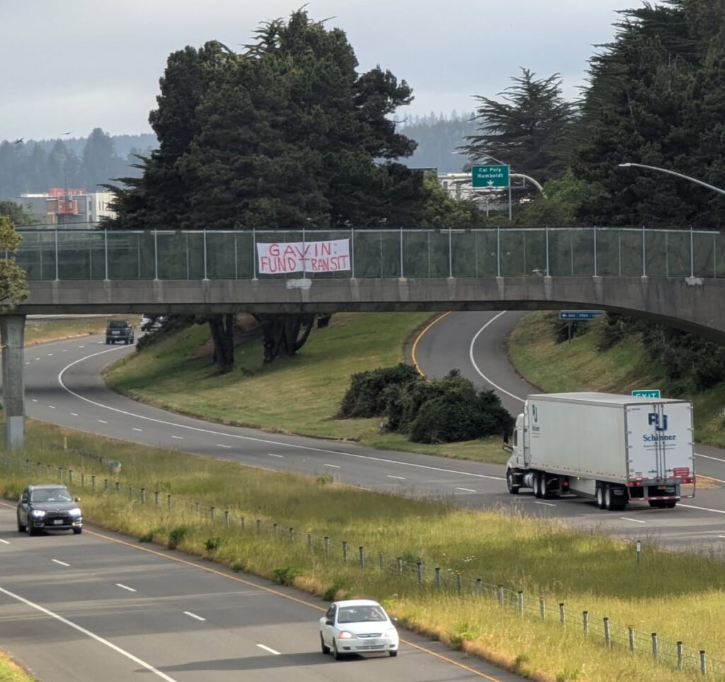 A wide divided highway is shown, with a broad grassy median and two lanes in each direction plus an exit ramp. A tractor trailer and several cars are visible. Over the highway, on an overpass, a hand-written banner reads: "Gavin: Fund Transit."