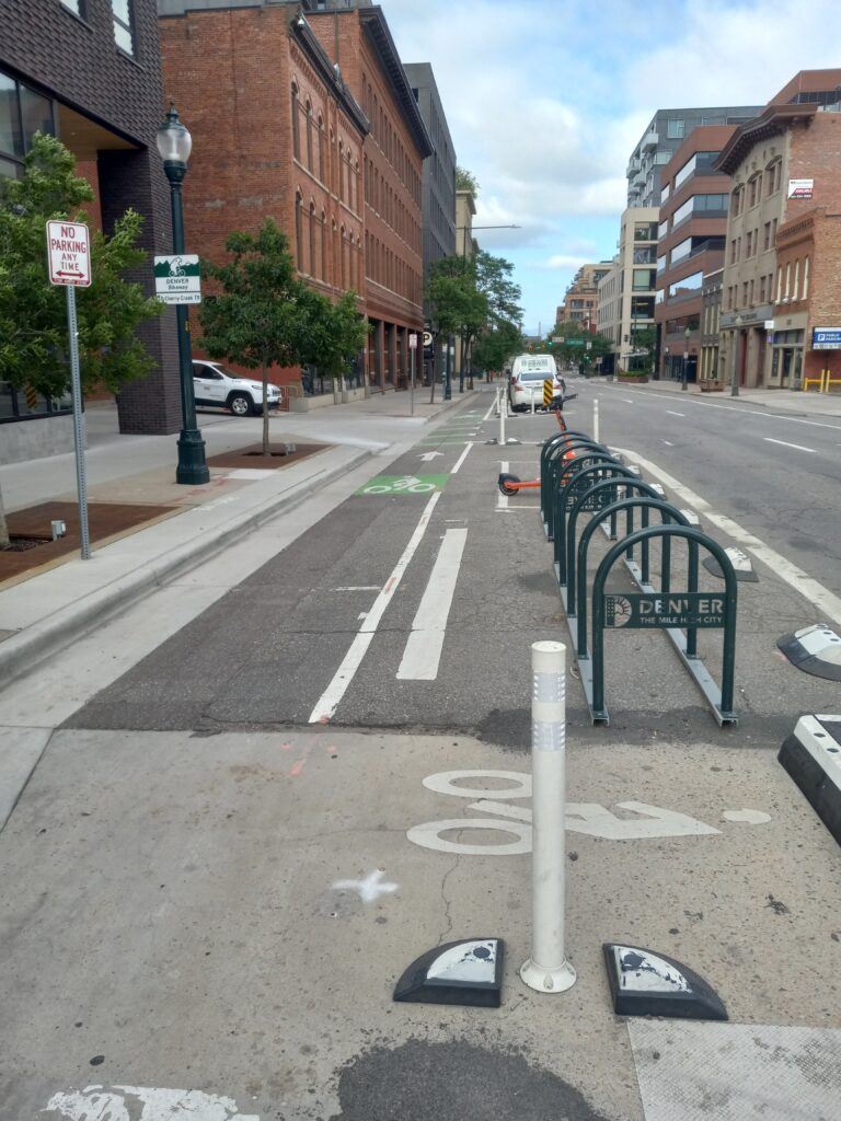 A city street between mostly brick, 3-story buildings has a row of metal inverted-U shaped bike racks in a converted parking spot, with low plastic humps and flexible posts between the bike parking and the street, and a bike lane between the bike parking and the sidewalk