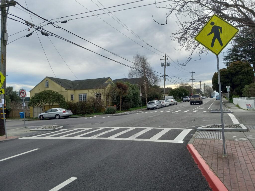 A crosswalk on a freshly paved two-lane street in the foreground, with older pavement in the background