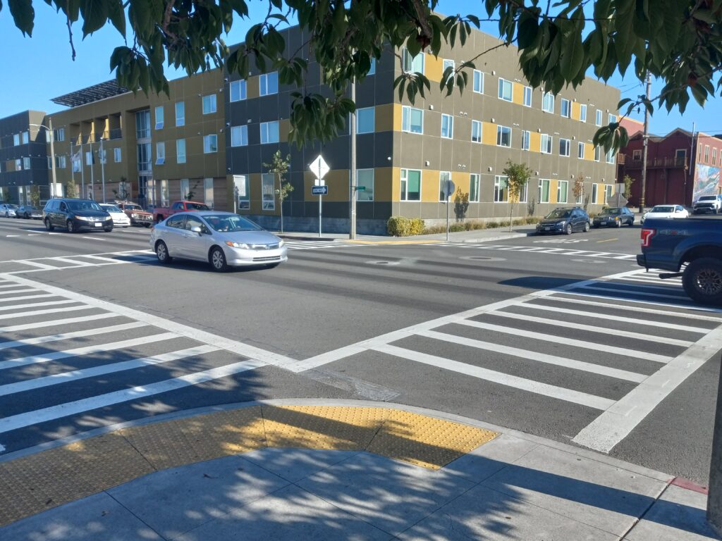 An intersection has four ladder-style crosswalks across a three-lane, one-way street and a smaller cross-street. There is a curb extension in the foreground and a three-story apartment building in the background.