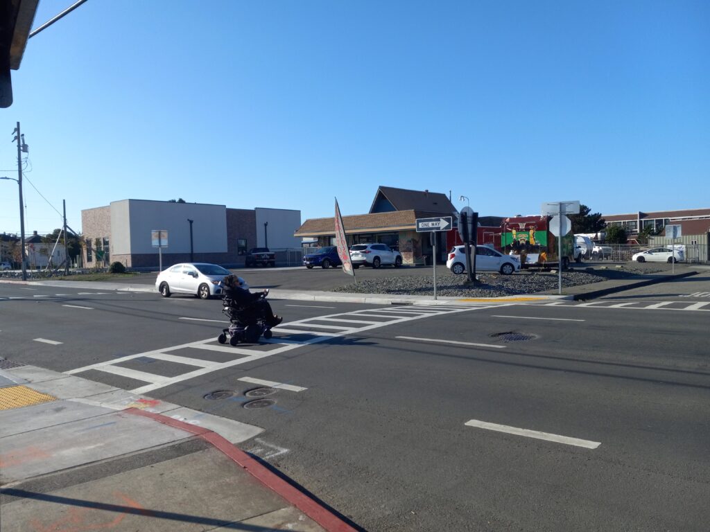 A person in a motorized wheelchair crosses a three-lane, one-way street in a marked crosswalk. A car is visible approaching in the far lane.