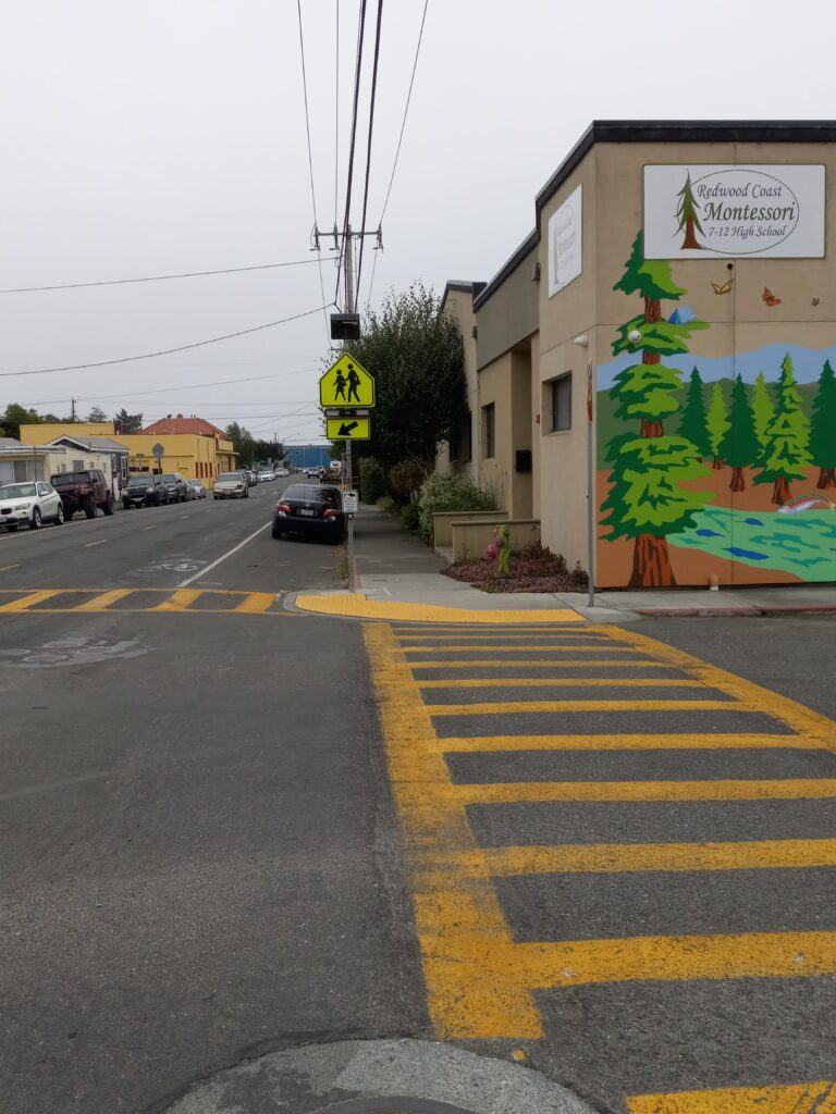 Two yellow crosswalks approach a corner with a yellow pedestrian sign and beacon on a corner with a low building. The building has a sign reading "Redwood Coast Montessori 7-12 High School."