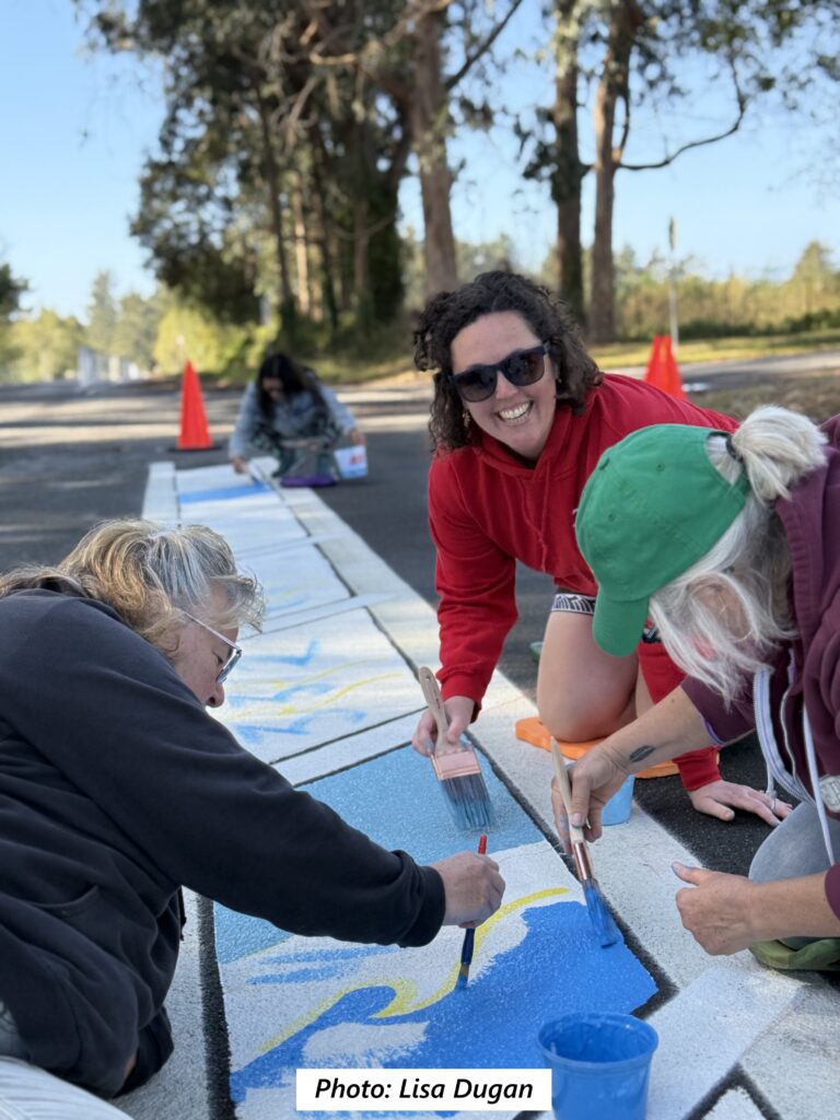 Three women are painting a strip of asphalt with blue and white paint. Two are bent over their work, while a third is smiling at the camera. Traffic cones and trees are visible in the background.