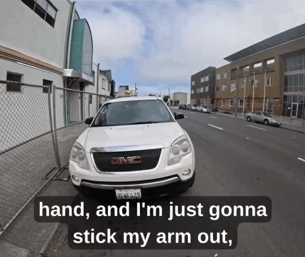 A point-of-view camera shot shows the front of a large white vehicle parked along a curb, with the sidewalk to the left blocked by a chain-link fence. To the right is a three-lane, one-way street.