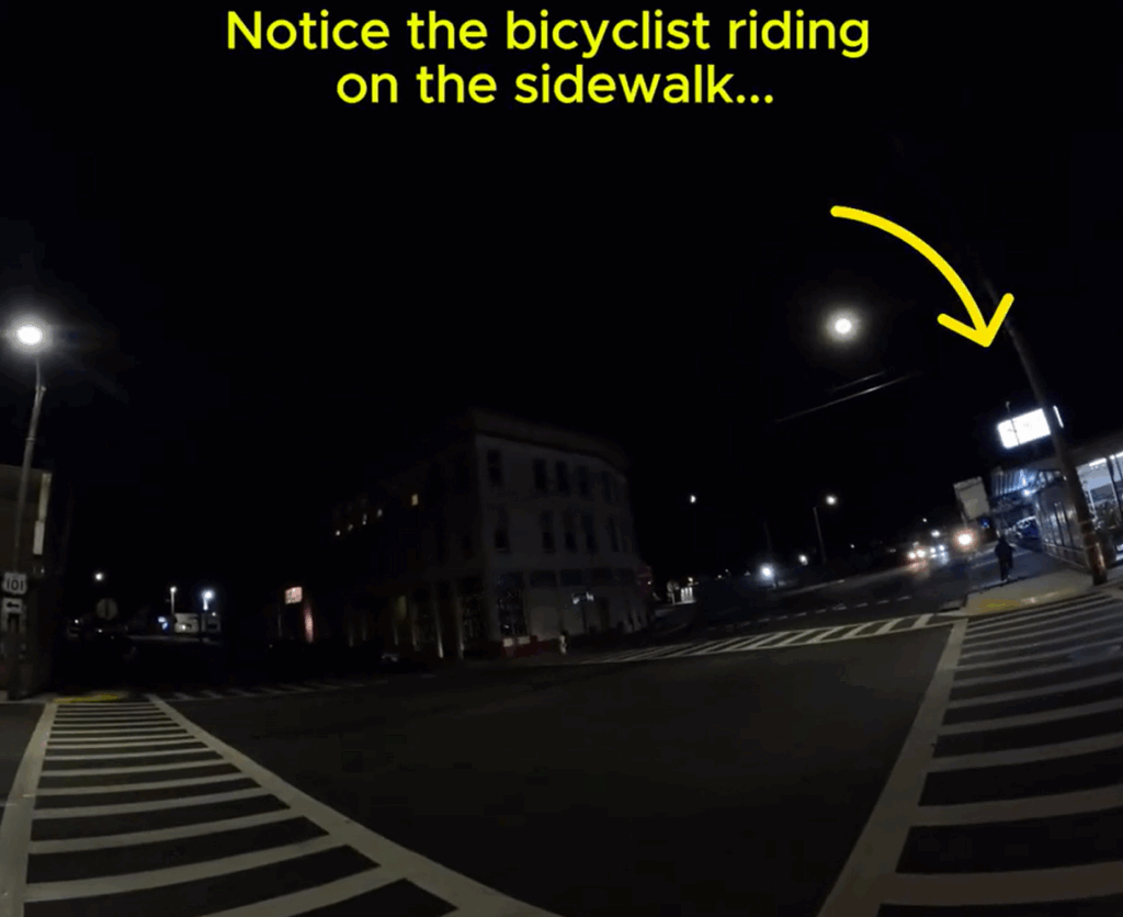 A nighttime intersection is shown, with visible crosswalks and a few streetlights, but otherwise very dark. A caption with an arrow reads: "Notice the bicyclist riding on the sidewalk..."