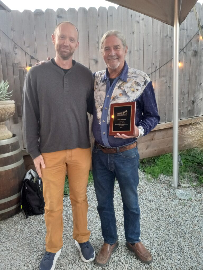Two men stand in front of a tall wooden fence. The man on the left is white, has short hair, and is wearing a gray sweater and orange pants. The man on the right is white, has long braided hair, and is wearing a multi-colored shirt and jeans and holding a plaque.