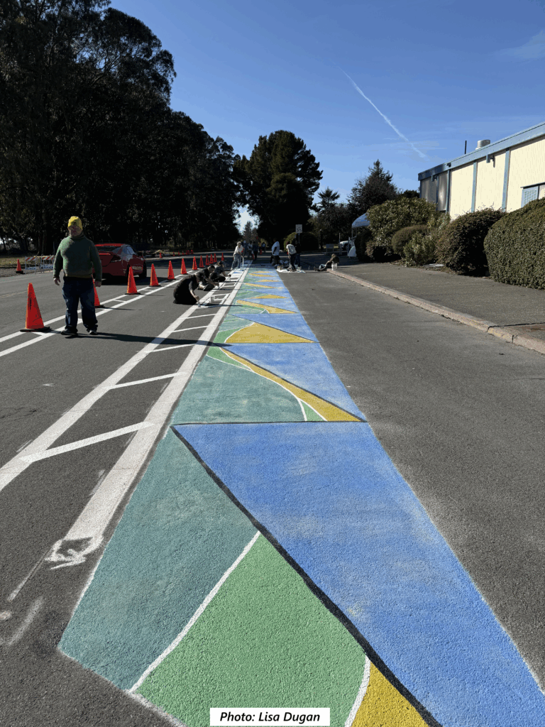 Looking down a wide street, a linear strip of blue, green, and yellow geometrically patterned mural recedes into the background, bordering a bike lane with white-striped buffers on both sides. People can be seen in the background working on the mural, protected by traffic cones.