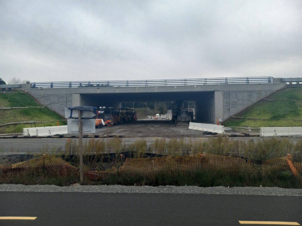 A highway underpass flanked by grassy slopes is shown. The underpass itself is packed dirt with many construction vehicles. Construction fencing and weeds are visible in the foreground.