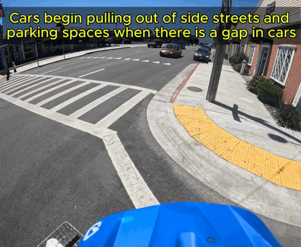 A view from on top of a bike helmet shows an intersection with yellow truncated domes on the sidewalk and a painted ladder-style crosswalk across three lanes of one-way traffic. Cars are approaching in all three lanes. A caption reads: Cars begin pulling out of side streets and parking spaces when there is a gap in cars."