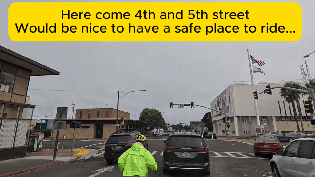 A person on a bike wearing a reflective neon green jacket and a helmet rides in the middle of a three-lane, one-way street, surrounded by cars approaching an intersection. A traffic light and office buildings are visible in the background. A caption reads "Here come 4th and 5th street. Would be nice to have a safe place to ride..."