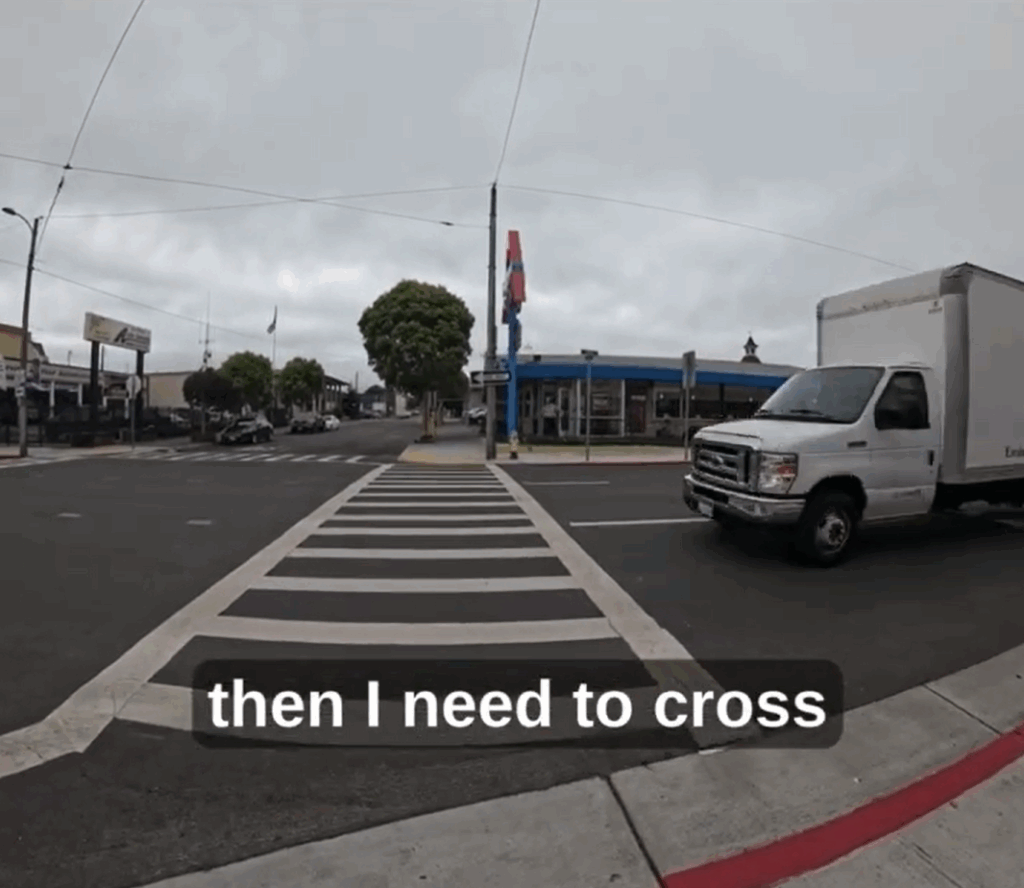 A still shot from a video shows a long crosswalk across a three-lane, one-way street, with a box truck approaching from the right. A restaurant is visible across the street. A caption reads "then I need to cross."