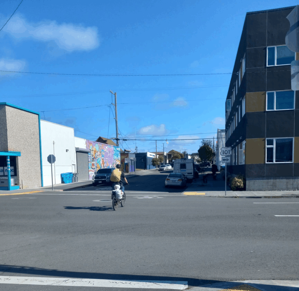 A bicyclist with a yellow backpack crosses a wide street. A one-story commercial building and a three-story apartment building are on the other side of the street.