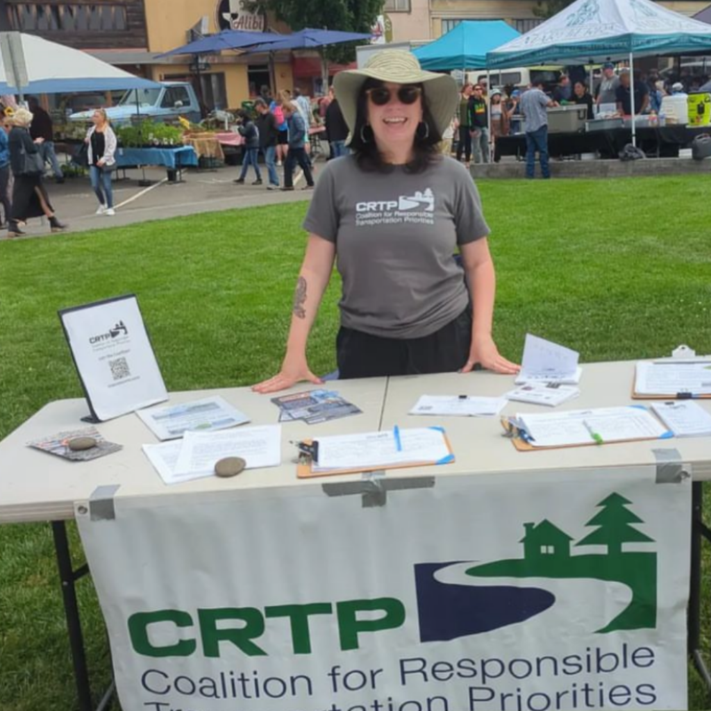 A young woman in a gray CRTP t-shirt, a wide-brimmed hat and sunglasses stands behind a table covered with piles of paper. Green grass and market stalls can be seen in the background, and the front of the table has a CRTP banner on it.