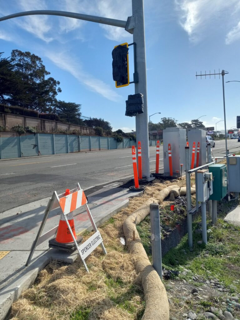 A new traffic signal still covered in black plastic is next to a wide street. Neon orange plastic bollards, cones and signs are clustered on the sidewalk.