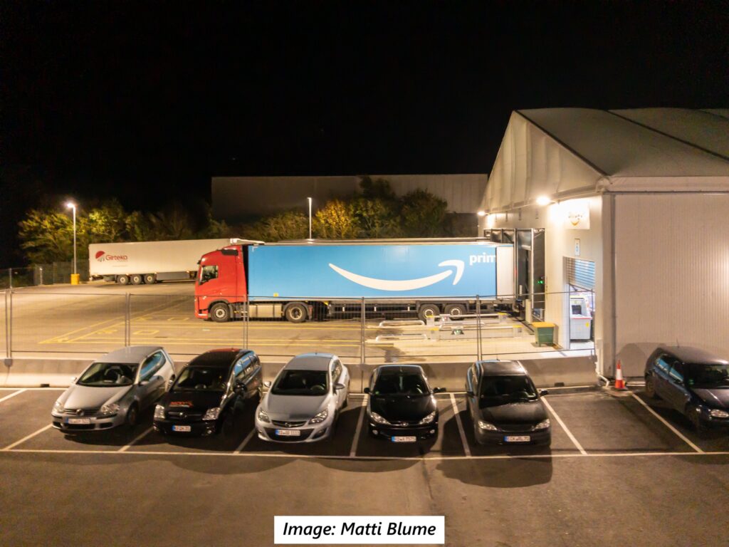 A row of parked cars in front of a chain link fence is in the foreground. In the background, a tractor trailer with the Amazon logo is backed up to a large warehouse.