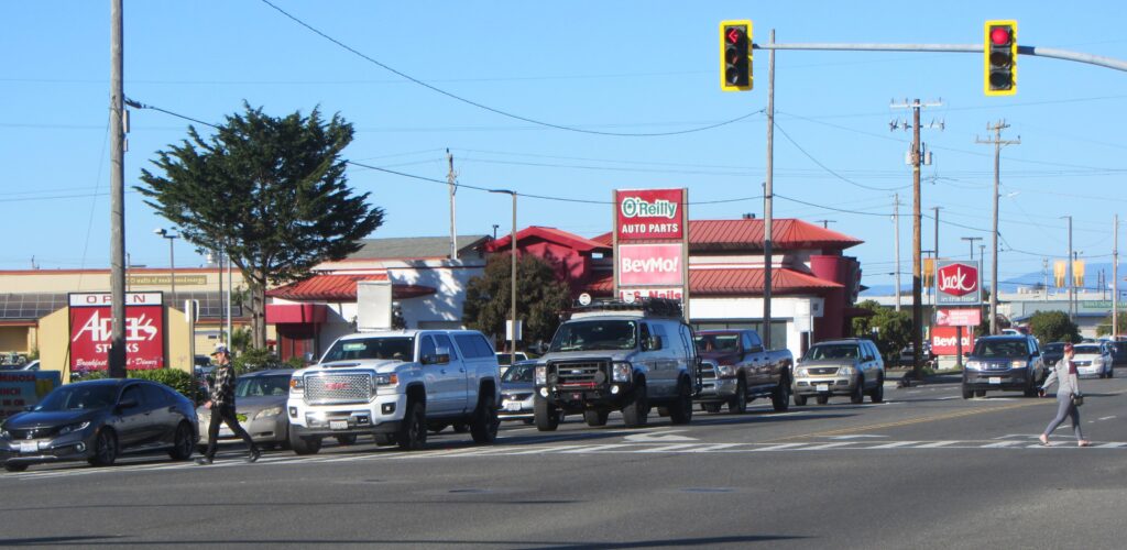 Two pedestrians are seen walking in a long crosswalk from the other side of a large intersection. Three lanes of cars face them, waiting at a red light.