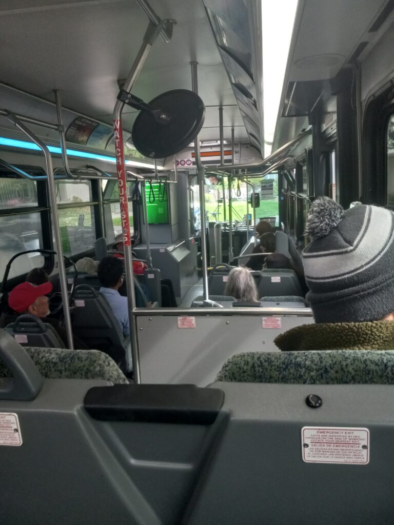 The picture is taken from inside the back of a bus, looking toward the front. Bus passengers with beanies and baseball hats are seated, with the backs of their heads visible.