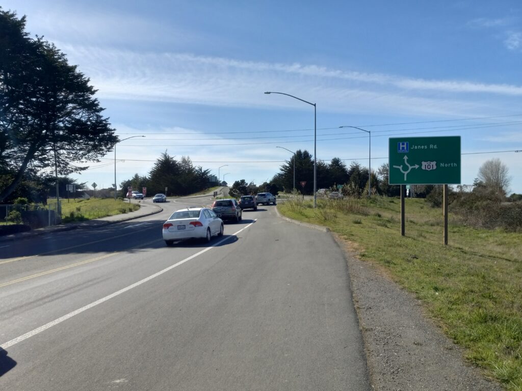 Cars line up entering a roundabout. A highway sign indicates the crossing of US 101.