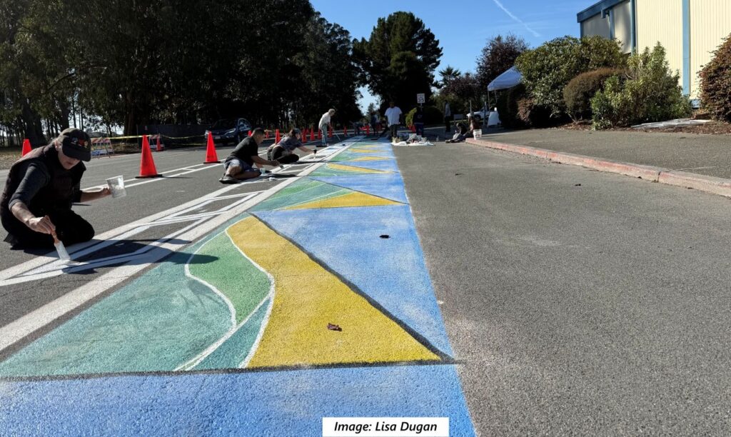 A strip of blue, yellow, and green painted mural stretches into the distance along a street. Several people are painting another strip of pavement alongside it.