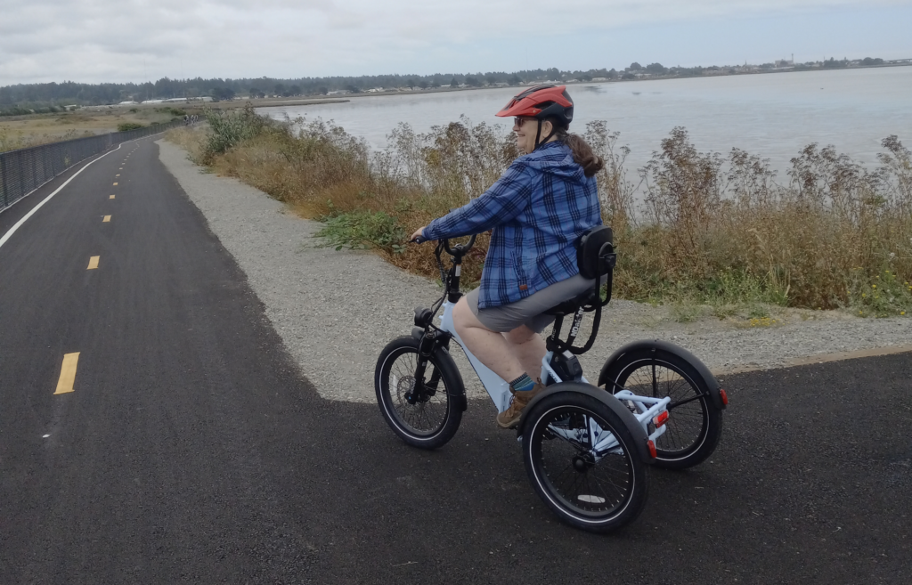 An older white woman wearing short, a blue plaid shirt and a red helmet rides a tricycle on a paved trail along a body of water.