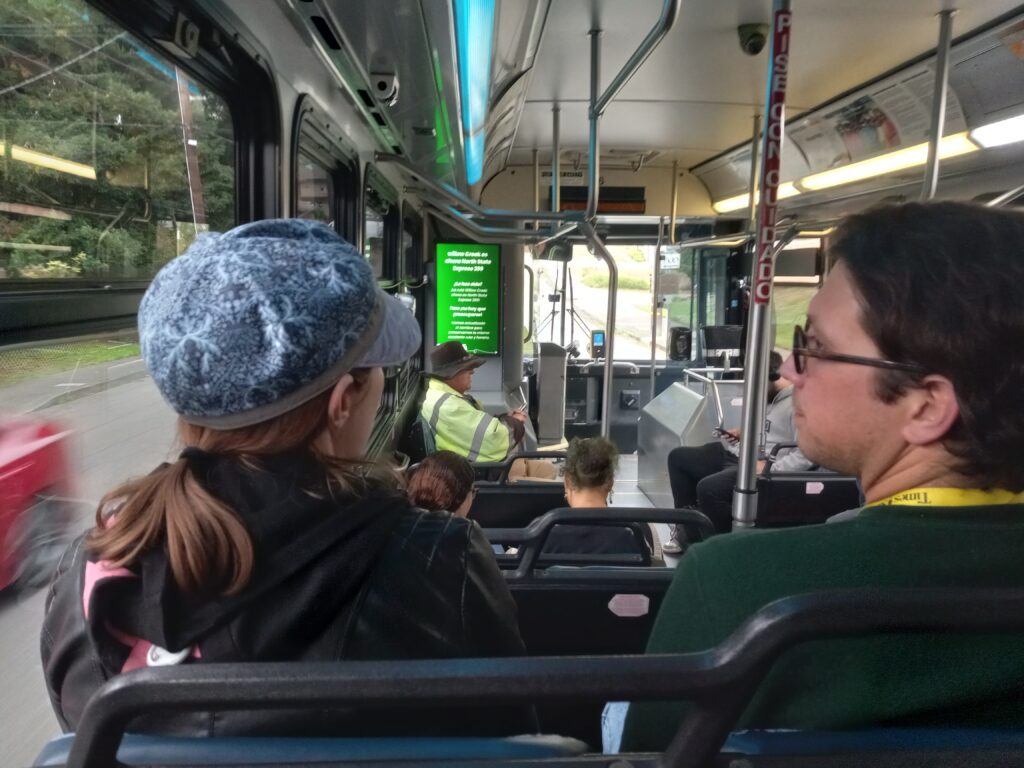 A white woman with long brown hair and a blue hat talks to a white man with glasses on a bus.