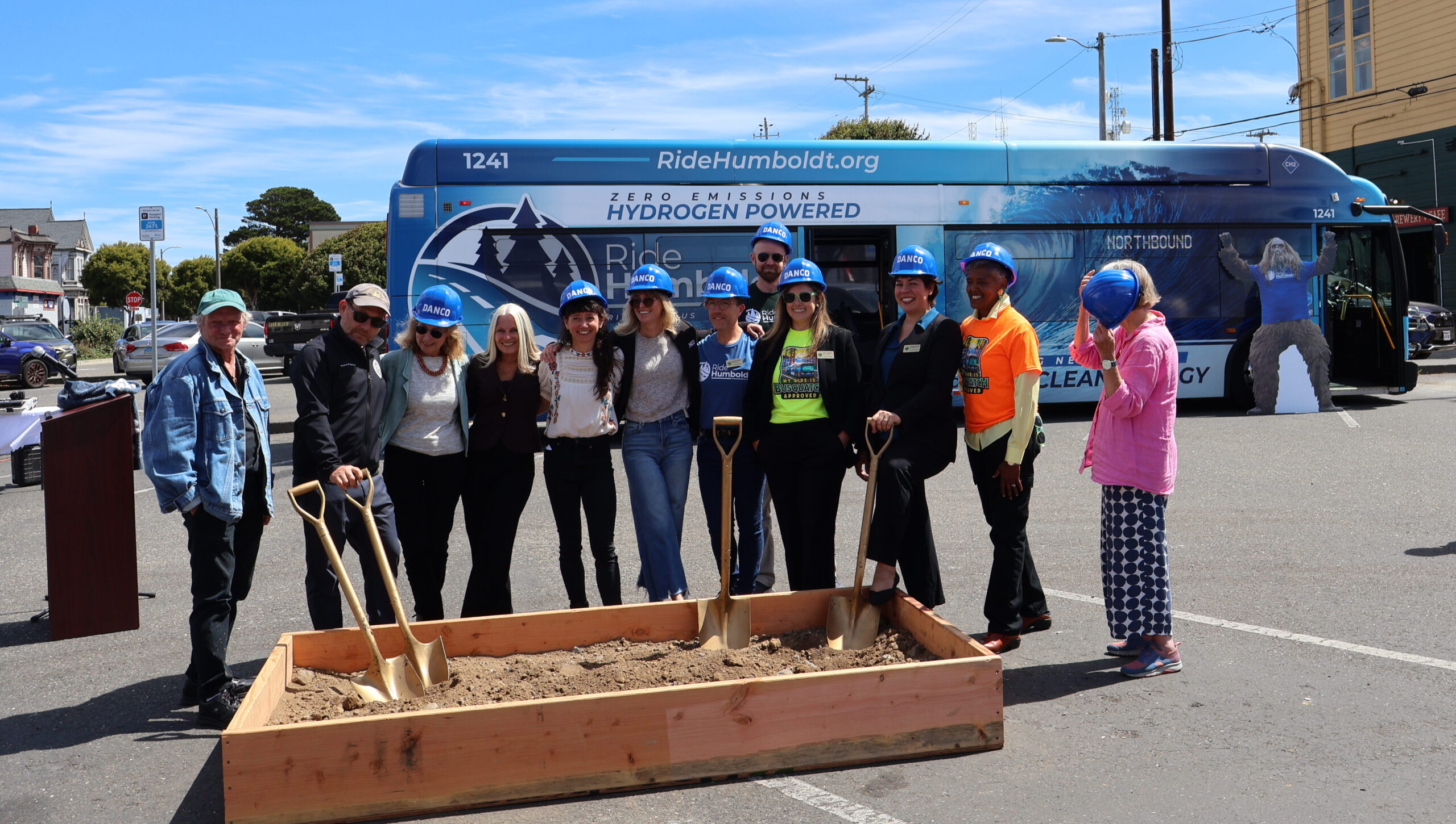 A group of people with hard hats and golden shovels stand behind a wooden box of dirt with a blue bus in the background.
