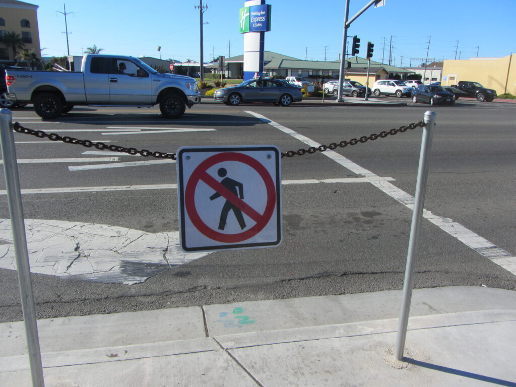 A sign with a pedestrian symbol in a circle with a line through it, hanging on a chain in front of a wide street.